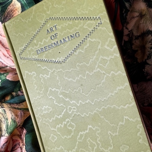 Puede incluir: Un libro de tapa dura titulado The Art of Dressmaking de Butterick con una cubierta de color verde oliva claro que presenta un patr&oacute;n de l&iacute;neas abstractas blancas y un borde en zigzag oscuro que rodea el t&iacute;tulo en forma de diamante centrado.