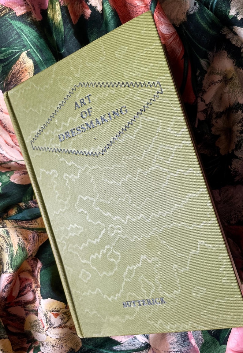 Puede incluir: Un libro de tapa dura titulado The Art of Dressmaking de Butterick con una cubierta de color verde oliva claro que presenta un patr&oacute;n de l&iacute;neas abstractas blancas y un borde en zigzag oscuro que rodea el t&iacute;tulo en forma de diamante centrado.