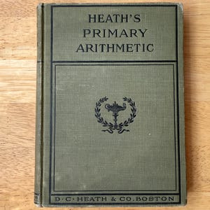 May include: A vintage hardcover textbook titled Heath's Primary Arithmetic with a sage green cloth cover. The front features black text and a central emblem of a lamp within a laurel wreath, with D.C. Heath & Co. Boston printed at the bottom.