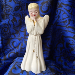 May include: A white porcelain figurine of an angel with blonde hair, wearing a white gown with gold trim and a gold rope necklace. The angel is praying with hands clasped together.