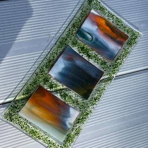 May include: A rectangular glass serving tray with a green speckled border. The tray holds three rectangular glass pieces with abstract designs in shades of orange, blue, and white. The tray is on a silver surface.