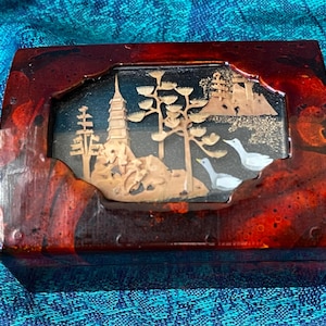 May include: A small, rectangular wooden box with a red and brown marbled finish. The box has a clear glass window that reveals a miniature scene of a pagoda, trees, and birds made of wood and other materials.