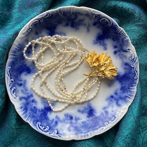 May include: A long necklace featuring small white freshwater pearls interspersed with tiny gold-toned beads, finished with a large, textured gold-toned metal floral pendant. The necklace is arranged in a coiled pattern on a white and blue ceramic plate.