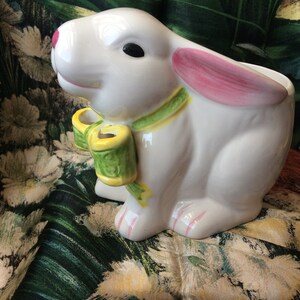 May include: A white ceramic bunny figurine with pink inner ears, black eyes, and a pink nose. It has a green and yellow bow around its neck. The bunny is a decorative item, possibly for Easter or spring.