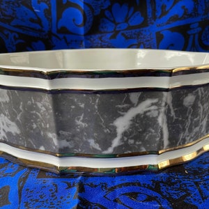 May include: A white ceramic bowl with a black and white marble design and gold trim. The bowl has a scalloped edge and is perfect for serving snacks or fruit.
