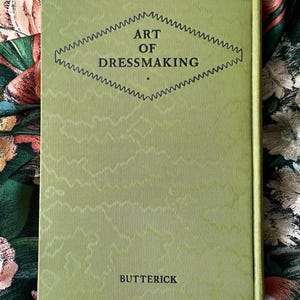 Puede incluir: Un libro de tapa dura titulado ART OF DRESSMAKING de BUTTERICK con una cubierta de color verde claro que presenta una textura acanalada vertical y un gr&aacute;fico central en forma de diamante delineado con un patr&oacute;n en zigzag. El texto est&aacute; en negro.