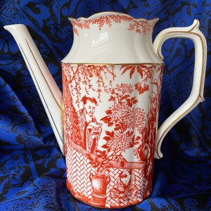 May include: A white porcelain teapot with a red and gold floral design. The teapot has a spout and a handle. The design features a Japanese-inspired scene with a figure in traditional clothing.