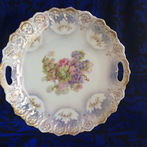 May include: A white porcelain serving tray with a scalloped edge and two handles. The tray features a floral design in the center with pink, purple, and green hues. The rim has a gold trim and decorative accents.