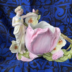 May include: A porcelain floral centerpiece featuring a woman in a flowing gown, leaning against a large pink flower. The flower has white and yellow accents, with green leaves and a branch handle. The woman has blonde hair and a blue jewel.