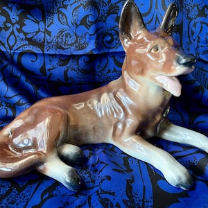 May include: A ceramic figurine of a brown and white German Shepherd dog lying down. The dog has a glossy finish and is posed with its head turned to the side.