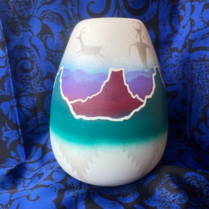 May include: A white ceramic vase with a hand-painted design of a mountain range, a purple sky, and two deer. The vase has a teal green base and a white top.