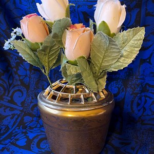 May include: A gold ceramic vase with a wire grid insert, holding a bouquet of artificial roses. The roses are white and peach in color, with green leaves.