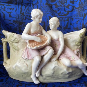 May include: A porcelain vase with two figures of women, one playing a musical instrument. The vase is decorated with floral details and has a cream and pink color scheme.