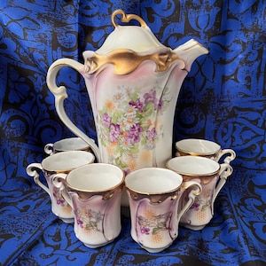 May include: A vintage porcelain tea set featuring a large teapot and six matching teacups. The set is decorated with floral patterns in shades of purple, white, and green, with gold trim. The teapot has a curved handle and a decorative lid.