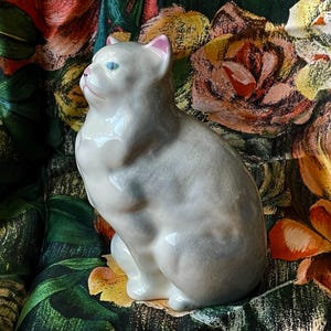 May include: A ceramic cat figurine in a seated position. The cat is light gray with pink inner ears and blue eyes. The background is a floral pattern with roses in shades of orange, pink, and yellow.