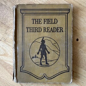 May include: The cover of a vintage book titled "The Field Third Reader". The cover is a tan color with a black silhouette of a Native American man in a circle. The circle is surrounded by mountains and trees.