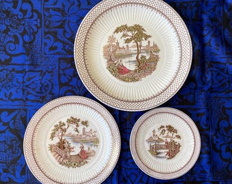 May include: A set of three white ceramic plates with a brown and gold floral design. The plates feature a scene of a couple walking through a garden with a bridge and a building in the background.
