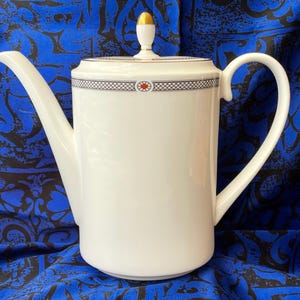 May include: A white porcelain coffee pot with a long spout and a curved handle. The lid features a gold-colored finial and a decorative band with a red accent. The pot is set against a blue patterned background.