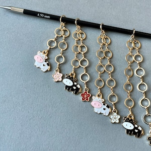 Könnte beinhalten: Fünf goldene Ketten mit Kuh-Anhängern und Blumenakzenten hängen an einer Stricknadel mit der Aufschrift "4,50 mm".