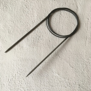 May include: A black circular knitting needle set against a textured, off-white background. The needles are dark grey with pointed tips, connected by a flexible black cable. The cable is coiled in a circular shape.