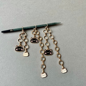 May include: Gold-toned earrings with a dangling design. Each earring features a series of gold rings connected to black cat charms with pink cheeks and white cat charms. A US size 7 knitting needle rests across the top.
