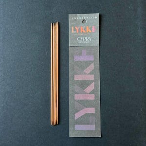 May include: Copper knitting needles from Lykke Crafts. The needles are a warm copper colour and are packaged with a grey paper label. The label has the Lykke Crafts logo and the words "CYPRA Copper Needles". The background is a dark grey.
