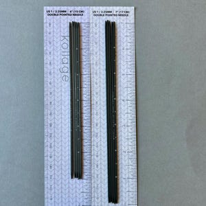 Può includere: Due set di ferri da maglia a doppia punta grigio scuro, uno da 15 cm e l'altro da 17 cm di lunghezza, sono esposti su uno sfondo in stile righello. Gli aghi sono etichettati "US 1 / 2.25MM" e "DOUBLE POINTED NEEDLE". È visibile anche il marchio "Kollage".