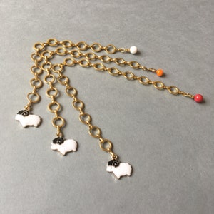 Könnte beinhalten: Goldfarbener Kettenschmuck mit weißen Schaf-Anhängern mit schwarzen Details. Die Ketten sind mit kleinen, farbigen Perlen in Weiß, Orange und Rot verziert. Der Schmuck wird vor einem grauen Hintergrund präsentiert.