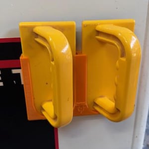 May include: Two bright yellow handles mounted on a white surface with a black and red stripe. The handles are rectangular with curved grips, designed for easy gripping and use. The handles are attached to a machine.