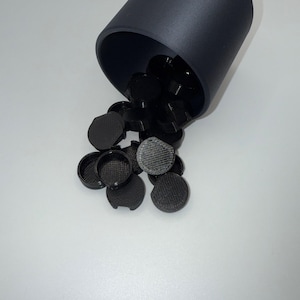 May include: A black cylindrical container tilted to the side, spilling out several small, round, black objects. The objects have a textured surface and a small lip around the edge. The background is a plain, light gray.