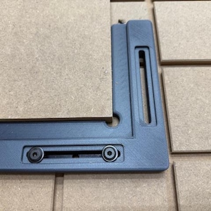 May include: A dark gray, right-angle woodworking jig with two black screws, holding a piece of light brown wood. The jig has a slot and a hole for alignment. The background features more light brown wood pieces.