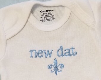 May include: White baby onesie with blue embroidered text that says "new dat" and a fleur-de-lis design. The onesie is made by Gerber and has the word "onesies" printed on the bottom.