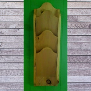 May include: A wooden wall-mounted organizer with three tiered pockets and a decorative top. The organizer is made of light-colored wood and is mounted on a green background. The organizer has a keyhole for hanging.