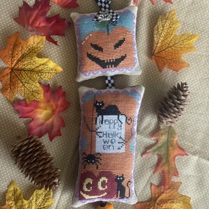 May include: A Halloween stitched ornament with a jack-o'-lantern design and the text "Happy Halloween". The ornament is orange, black, and white, with a checkered ribbon. Decorative autumn leaves and pine cones surround the ornament.