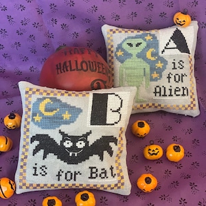 May include: Two handmade Halloween-themed cushion covers with embroidered letters and images. The first cushion cover is light grey with a black bat and the text "B is for Bat". The second cushion cover is light grey with a green alien and the text "A is for Alien".