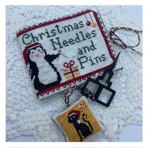 May include: A cross-stitched needle case with the text "Christmas Needles and Pins" and a penguin design. A small, square ornament with a black dog wearing a Santa hat is also visible, along with black scissors and a red and green cord.