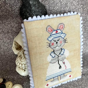 Puede incluir: Un bordado en punto de cruz cuadrado sobre tela color canela que muestra un ratón gris vestido de enfermero con gorro blanco, ribetes azules y un estetoscopio. La pieza está bordeada con pompones blancos y tiene una cruz roja.