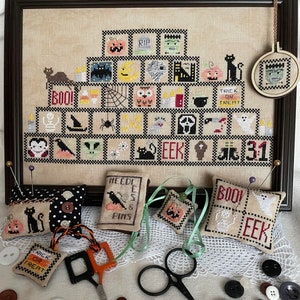 May include: A framed cross-stitch design with a Halloween theme. The design features a pyramid of squares with various Halloween icons, including pumpkins, bats, ghosts, and spiders. The frame is brown wood and the fabric is a beige linen. There are also several small needlepoint cushions with Halloween themes, including a black cat, a pumpkin, and a ghost.
