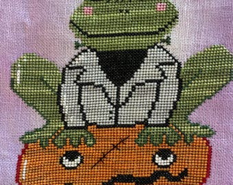 Könnte beinhalten: Kreuzstichmuster einer grünen Froschfigur in einem weißen Jackett mit schwarzem Kragen, der auf einem orangefarbenen Kürbis mit einem Kürbisgesicht sitzt. Ein kleines rotes Herz schwebt darüber. Der Hintergrund ist hellviolett.