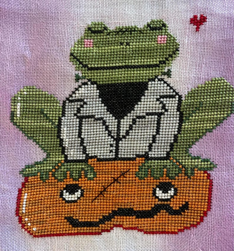 May include: Cross-stitch design of a green frog in a white jacket with a black collar, perched on an orange pumpkin with a jack-o'-lantern face. A small red heart floats above. The background is a light purple.