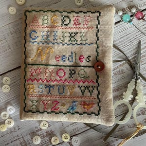 May include: A beige needlepoint alphabet chart with a brown button and a tassel. The chart is surrounded by white buttons on a white wooden surface.