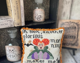 Puede incluir: Un cojín bordado con ribete naranja presenta una bruja con cabello rojo y el texto "Oh look, another glorious morning makes me sick." Una botella de vidrio etiquetada "Snake Venom" es visible, junto con otros artículos temáticos de boticario.