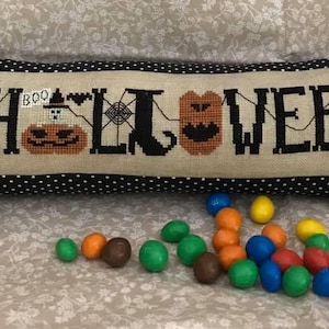 May include: A cylindrical candy-shaped cushion featuring a black and white polka dot fabric with purple and white ribbons at the ends. The centre panel displays cross-stitched text reading HALLOWEEN with pumpkin, spiderweb, and candy corn motifs.