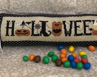 May include: A cylindrical candy-shaped pillow featuring a black and white polka dot fabric with purple and white ribbons at the ends. The center panel displays cross-stitched text reading HALLOWEEN with pumpkin, spiderweb, and candy corn motifs.