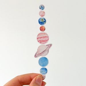 May include: A vertical sticker sheet with watercolour illustrations of the planets. The planets are in shades of blue, pink, red, and orange, with Saturn's rings visible. The sticker sheet is held by a hand, against a white background.