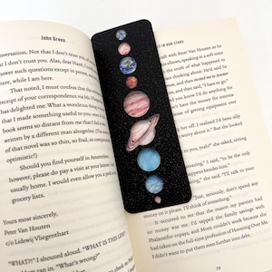 May include: Black bookmark with a vibrant illustration of the planets in our solar system set against a starry backdrop. The bookmark is placed in a book. A distinctive and educational accessory for book lovers.