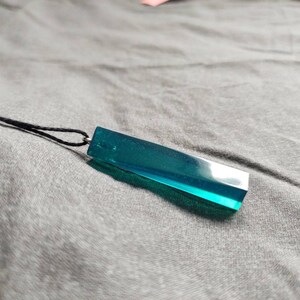 May include: A teal-coloured rectangular pendant necklace. The pendant is made of a translucent material and is suspended from a black cord. The necklace is on a grey surface.