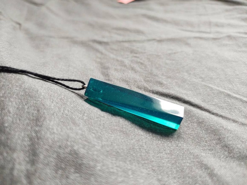 May include: A teal-coloured rectangular pendant necklace. The pendant is made of a translucent material and is suspended from a black cord. The necklace is on a grey surface.