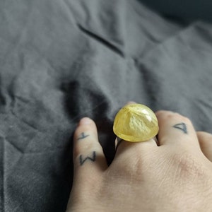 May include: A handmade ring featuring a large, rounded, yellow resin stone with a floral pattern. The ring is set on a silver band. The ring is on a hand with tattoos. The resin stone is approximately 2.5 cm in diameter.