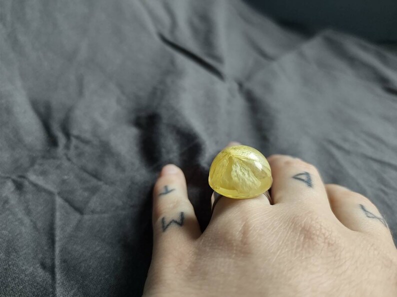 May include: A handmade ring featuring a large, rounded, yellow resin stone with a floral pattern. The ring is set on a silver band. The ring is on a hand with tattoos. The resin stone is approximately 2.5 cm in diameter.